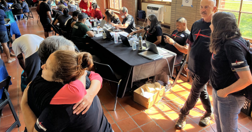 Image of two women hugging each other while surrounded in a room full of people working and helping people