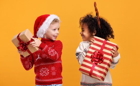 Happy excited multiracial children in Christmas outfit holding Xmas gifts while standing isolated over yellow studio wall background, two little boys of different races getting New Year presents