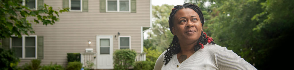 A person in foreground looking away from the camera with a slight smile. A house is in the background.
