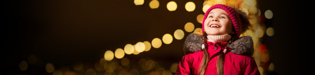 Child in winter clothes smiling and looking up at holiday lights
